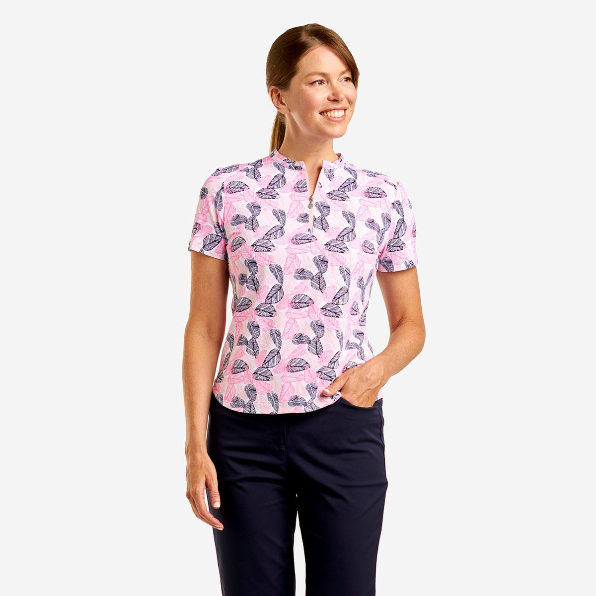 A woman modeling a short-sleeve collared polo-style top with a pink base and an allover abstract print in dark blue and white, paired with dark navy pants against a light gray studio background.