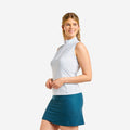 A woman modeling a sleeveless white athletic top with a high mock neck and a teal blue skirt, photographed against a solid light gray background.